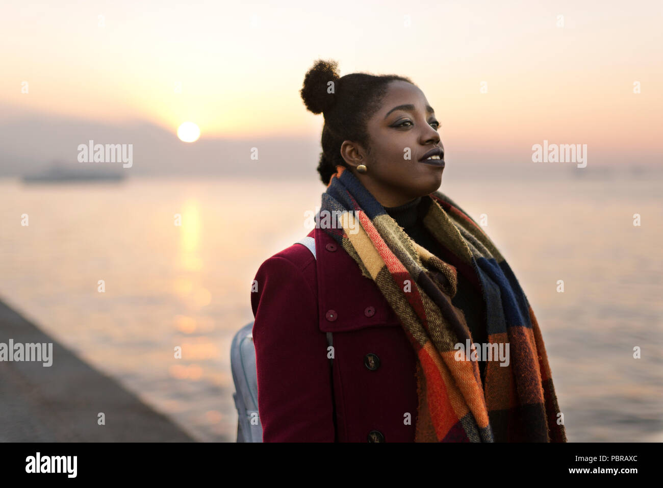 Portrait of a 25 years old African woman with a red coat and weft near to the waters edge with a sunset. Stock Photo