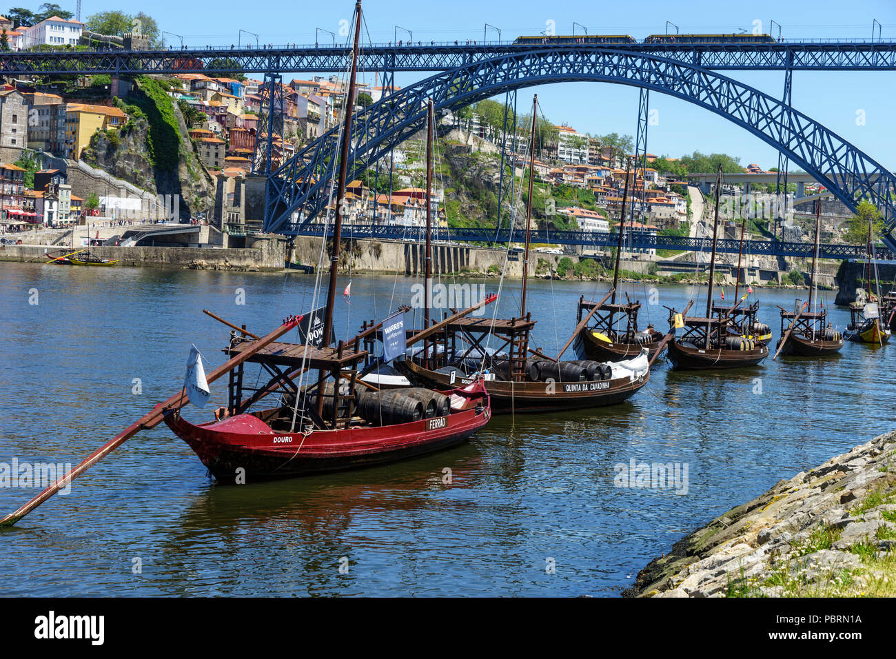 Traditional rabelo cargo boats moored off the bank of the River Douro with the double-deck metal arch bridge Luis 1 bridge in the background Porto Stock Photo