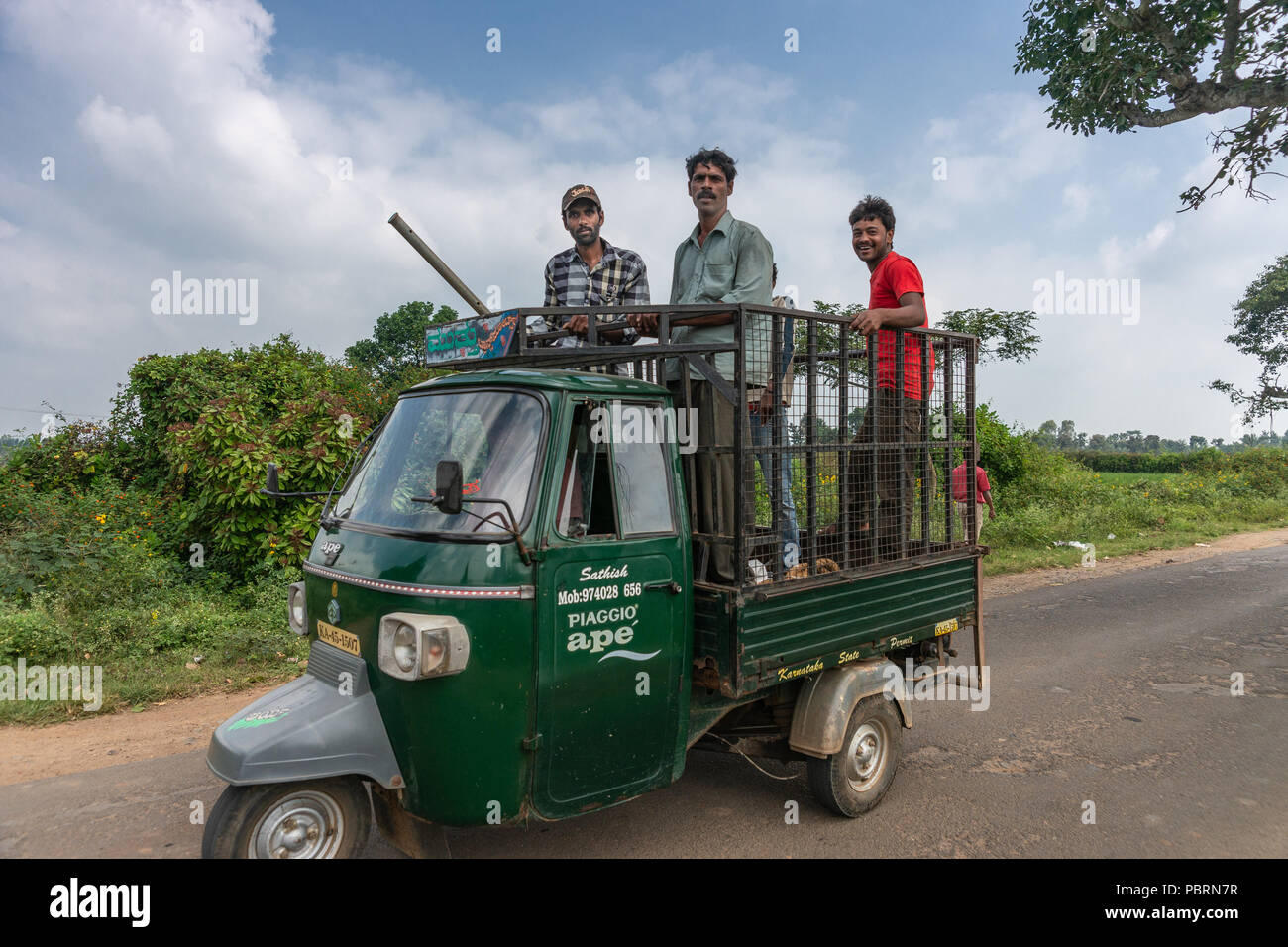 Belathur, Karnataka, India - November 1, 2013: Dark green tricycle car brings four farm workers, smiling men, standing on loading area of car to the f Stock Photo