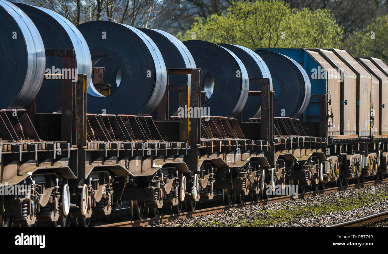 The back of a long and heavy rail freight train of wagons and flatbed trucks carrying large steel coils from a steelworks Stock Photo