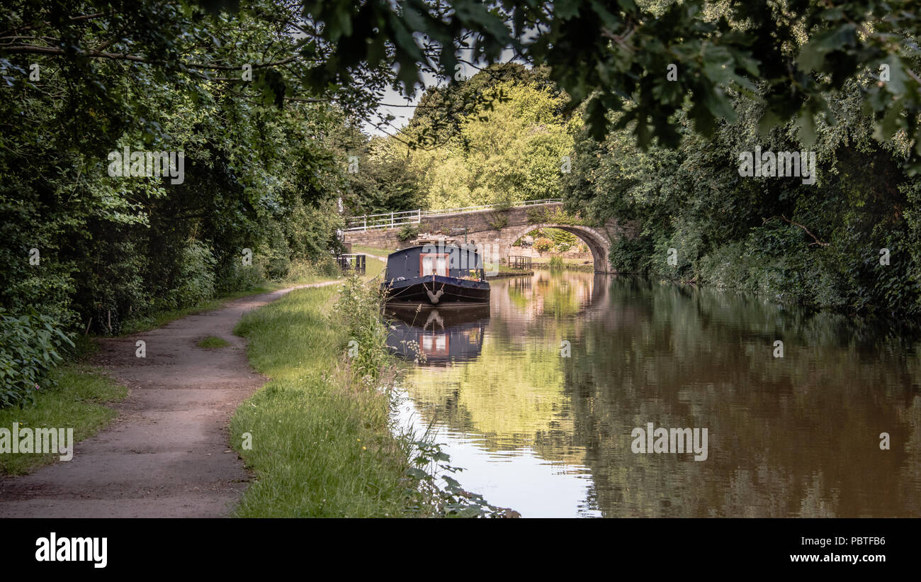 A peaceful scene looking down a canal towpath. There is a narrow boat moored up in front of an old road bridge crossing the water.. The scene is set i Stock Photo