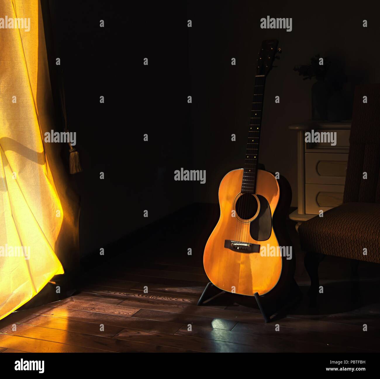 Acoustic Spanish guitar on a stand in the moody shadows of a dark room with bright light coming in from behind a curtain Stock Photo
