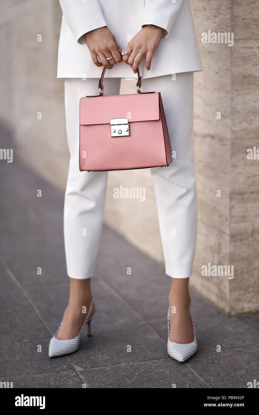 Beautiful girl is posing with a coral bag on the wall background outdoors. She wears a white pantsuit with light shoes. Closeup vertical photo. Stock Photo