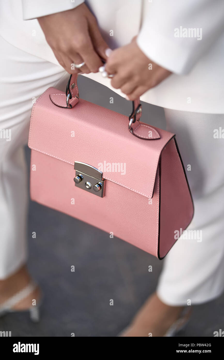 Nice girl is posing with a coral bag on the wall background outdoors. She wears a white pantsuit with light shoes. Closeup vertical photo. Stock Photo