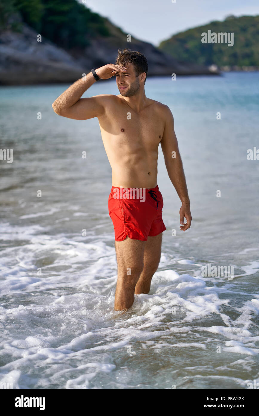 Athletic tanned man stands in the sea waves on the sunny nature background.  He wears a