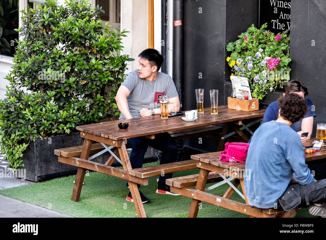 London, UK - June 22, 2018: People sitting on sidewalk street in England, by Sawyers Arms pub drinking pint of beer in glass with one young asian man, Stock Photo