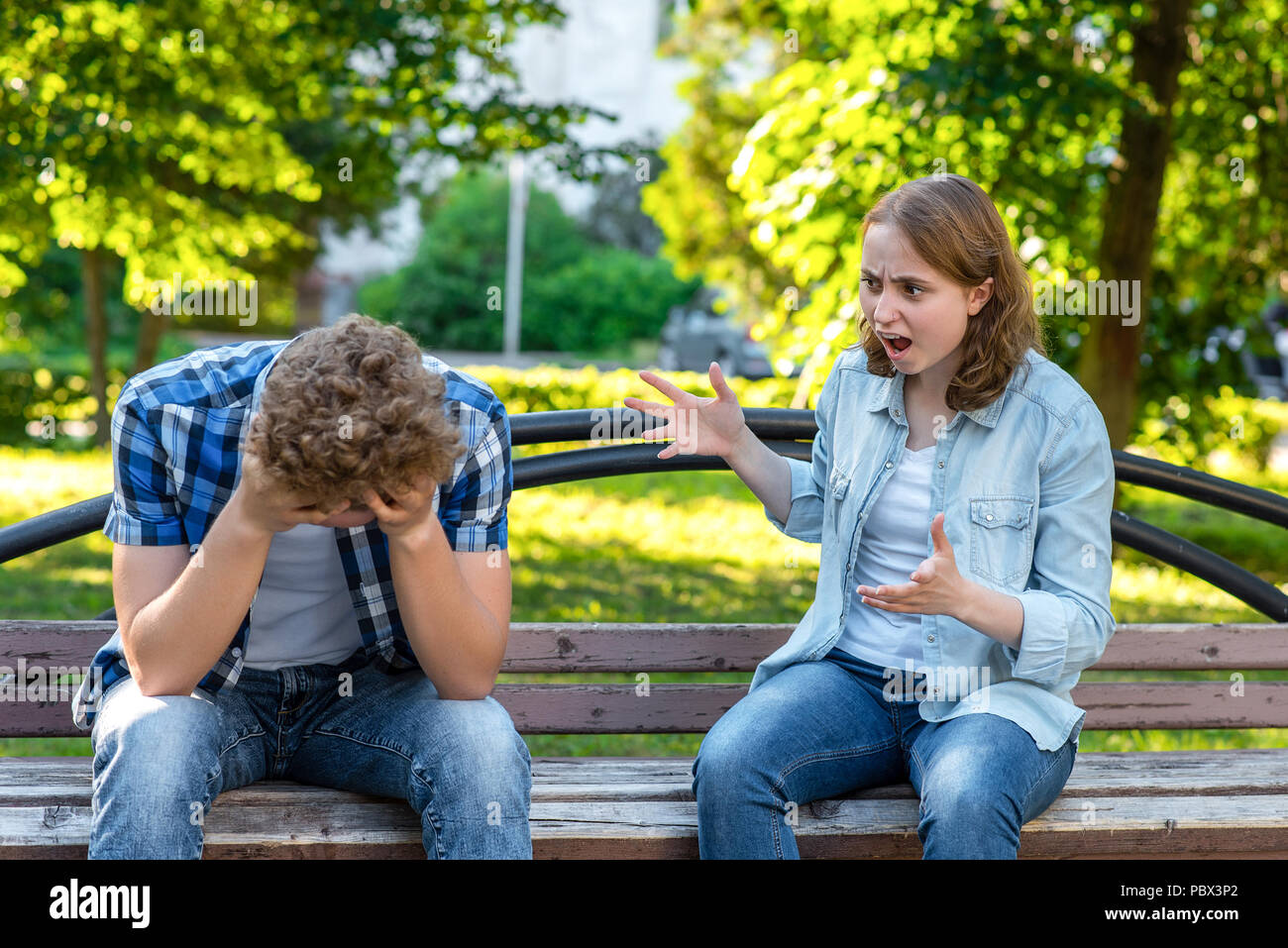 The girl screams at young man. Summer in park on a bench. The guy is crying and sad, clasping his head in his hands. Scandal in family. The concept of aggression and the problem in relationship. Stock Photo