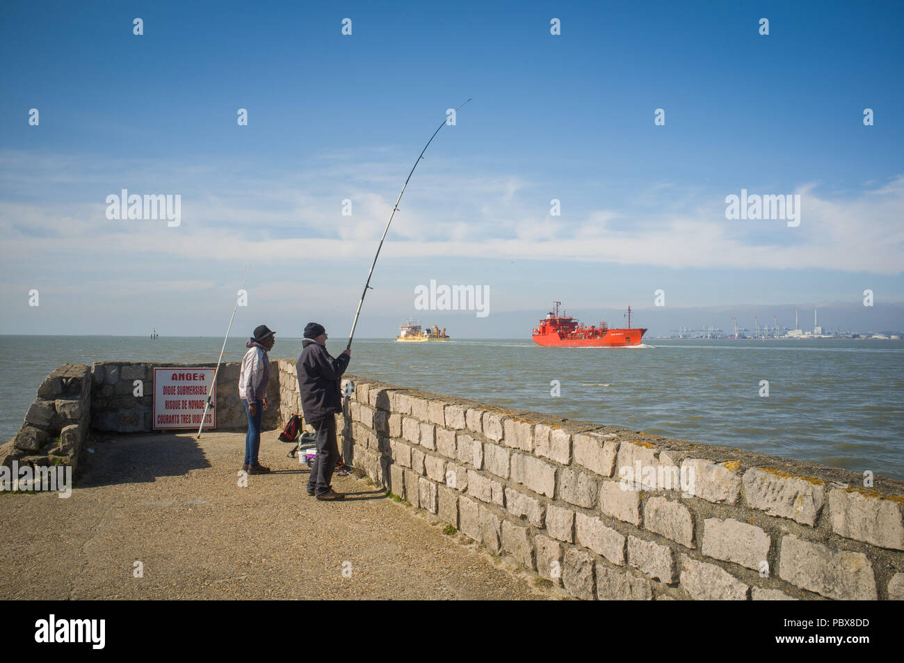 Two men fish from the quay at Honfleur as oil tankers pass in the Seine Estuary with Le Havre in the distance Stock Photo