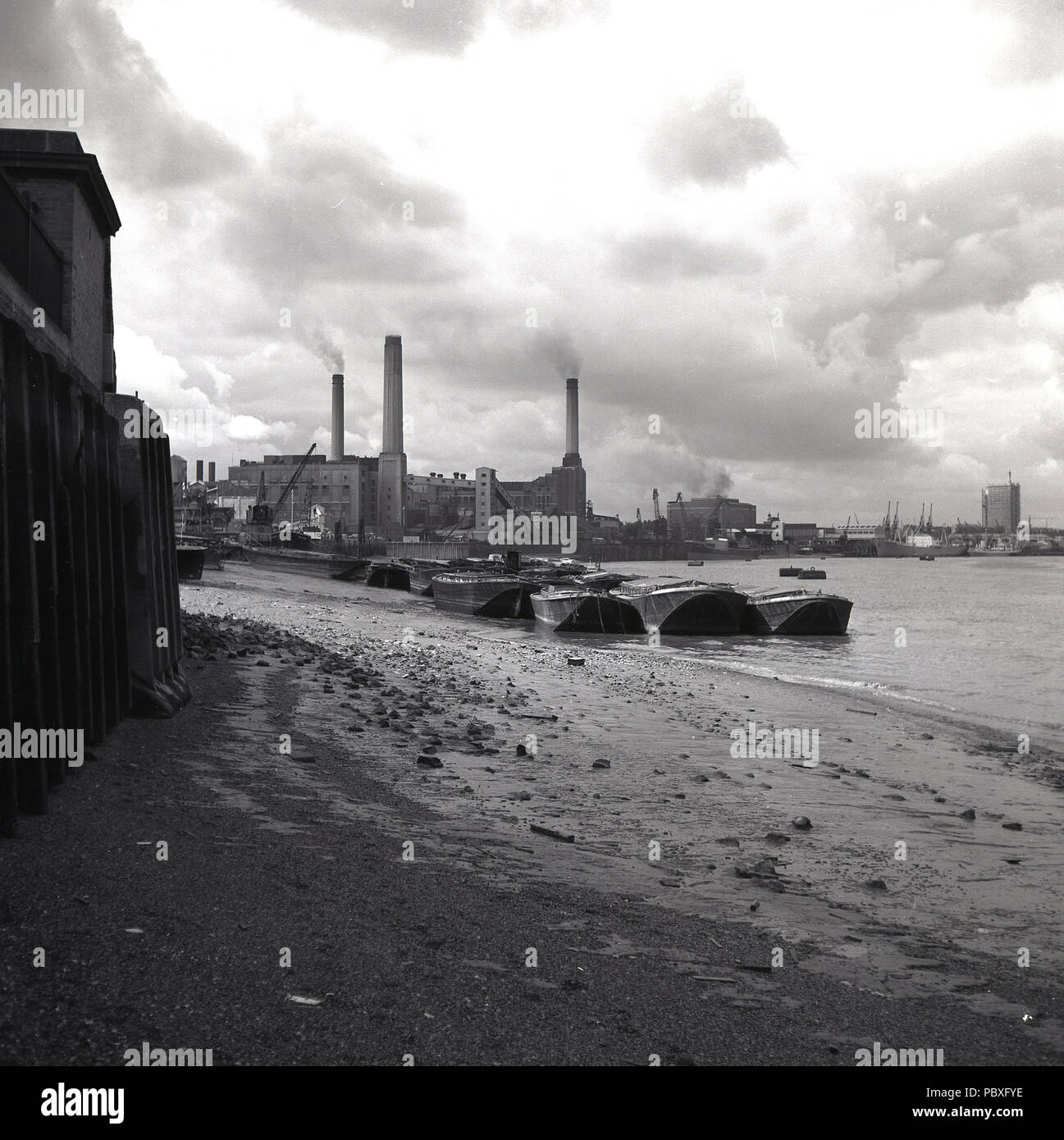 1950s, historical, the tide is out as barges sit on the south-bank of the river Thames by a small pebbly beach with a view  from the east of a working Deptford Power Station in the distance, London, England, UK. Designed by Sebastian de Ferranti, Deptford is know as the world's first central power station. Coal fired, it was unprecedented in both its scale and high voltage and it continued operating until the late 1960s. Stock Photo