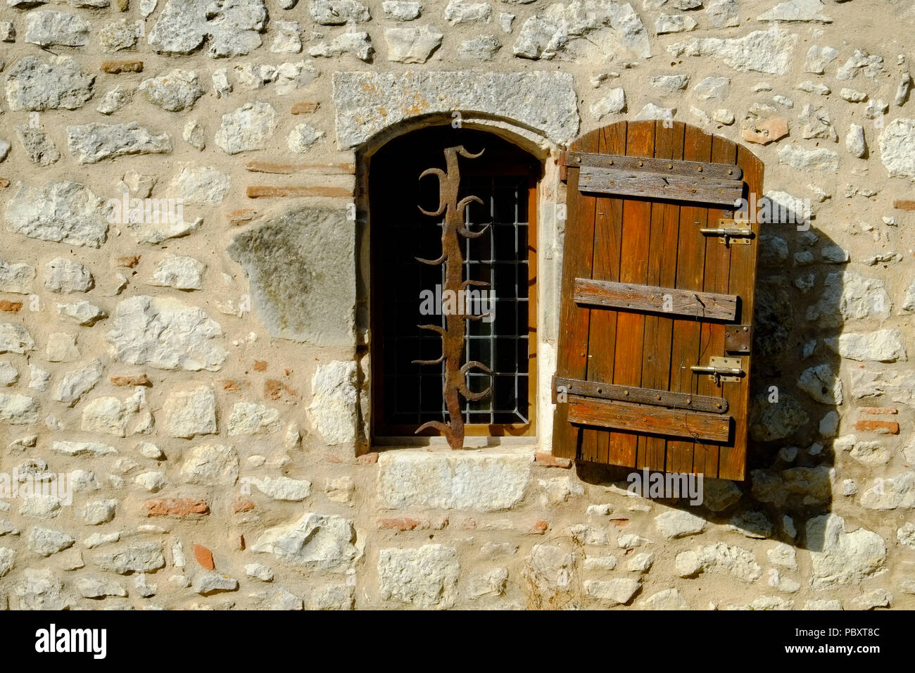 A rustic shuttered window with ornate security bars set in a stone wall in Pujols, Lot-et-Garonne, France. This historic village is a member of 'Les Plus Beaux Villages de France' association. Stock Photo