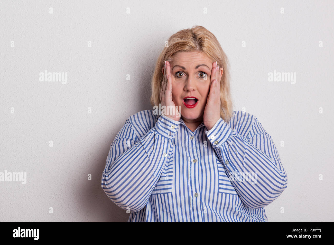 Portrait of a scared overweight woman in studio, hands on cheeks. Stock Photo
