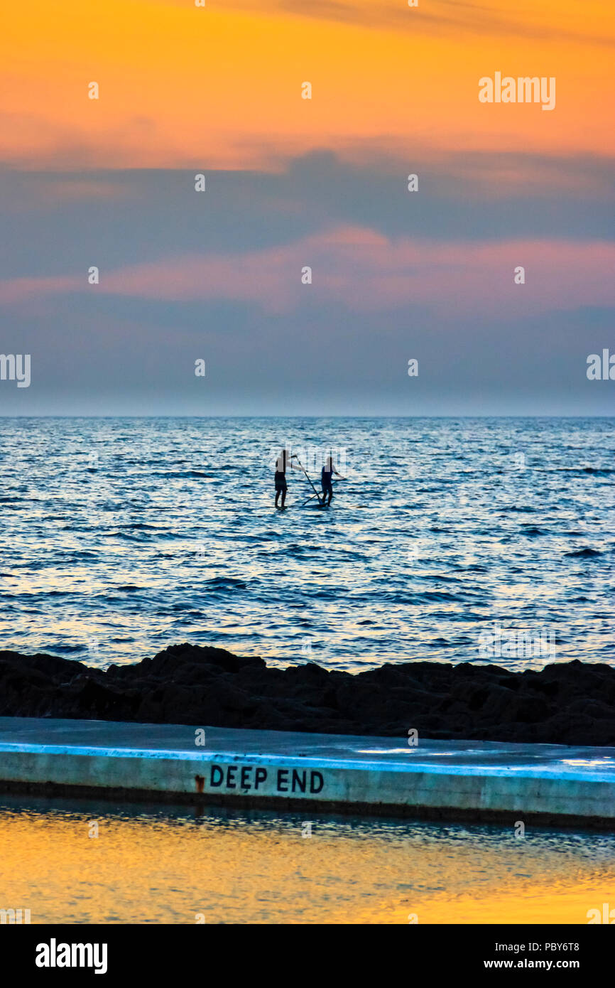 Two paddleboarders silhouetted against the sunset at Westward Ho!, Devon, UK, the seawater swiiming pool in the foreground Stock Photo