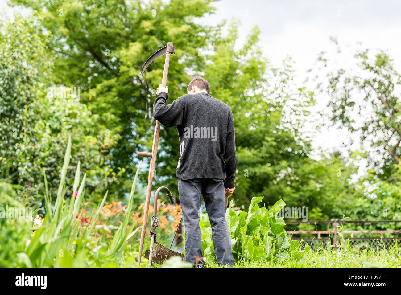 Back of young man farmer in garden standing with sickle scythe tool in green summer in Ukraine by well faucet Stock Photo