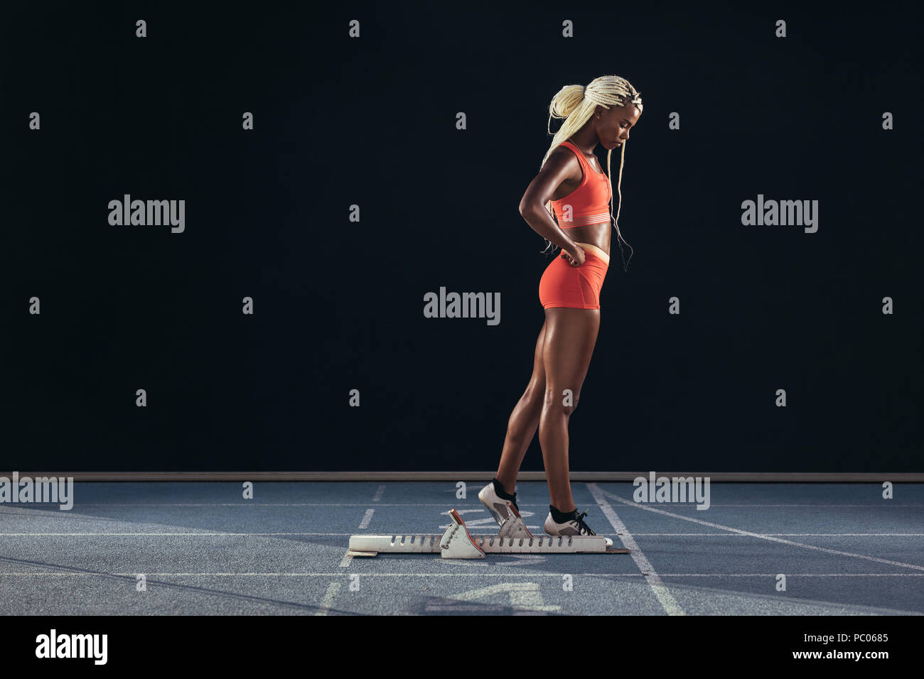 Female sprinter standing beside a starting block on running track on a black background. Side view of a female runner standing at the start line on an Stock Photo