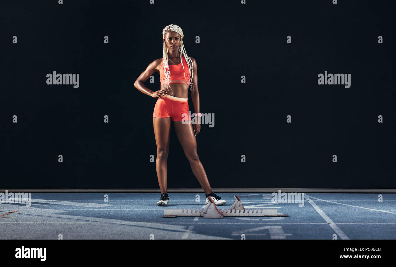 Female athlete standing beside a starting block on running track on a black background. Female runner standing at the start line on a running track wi Stock Photo