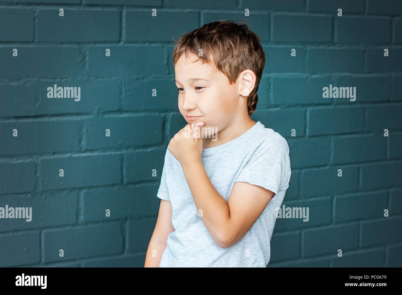 Boy 8 years old with freckles looking into the distance, thinking and scratching his head against the gray brick wall Stock Photo