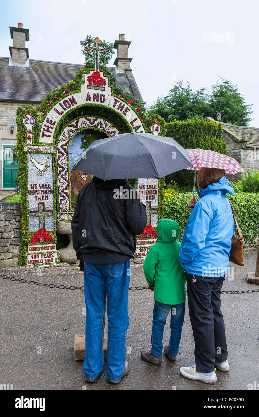 A family stand in the rain under umbrellas, looking at Hands Well, Tissington Well Dressing in the village of Tissington, Derbyshire, England, UK Stock Photo