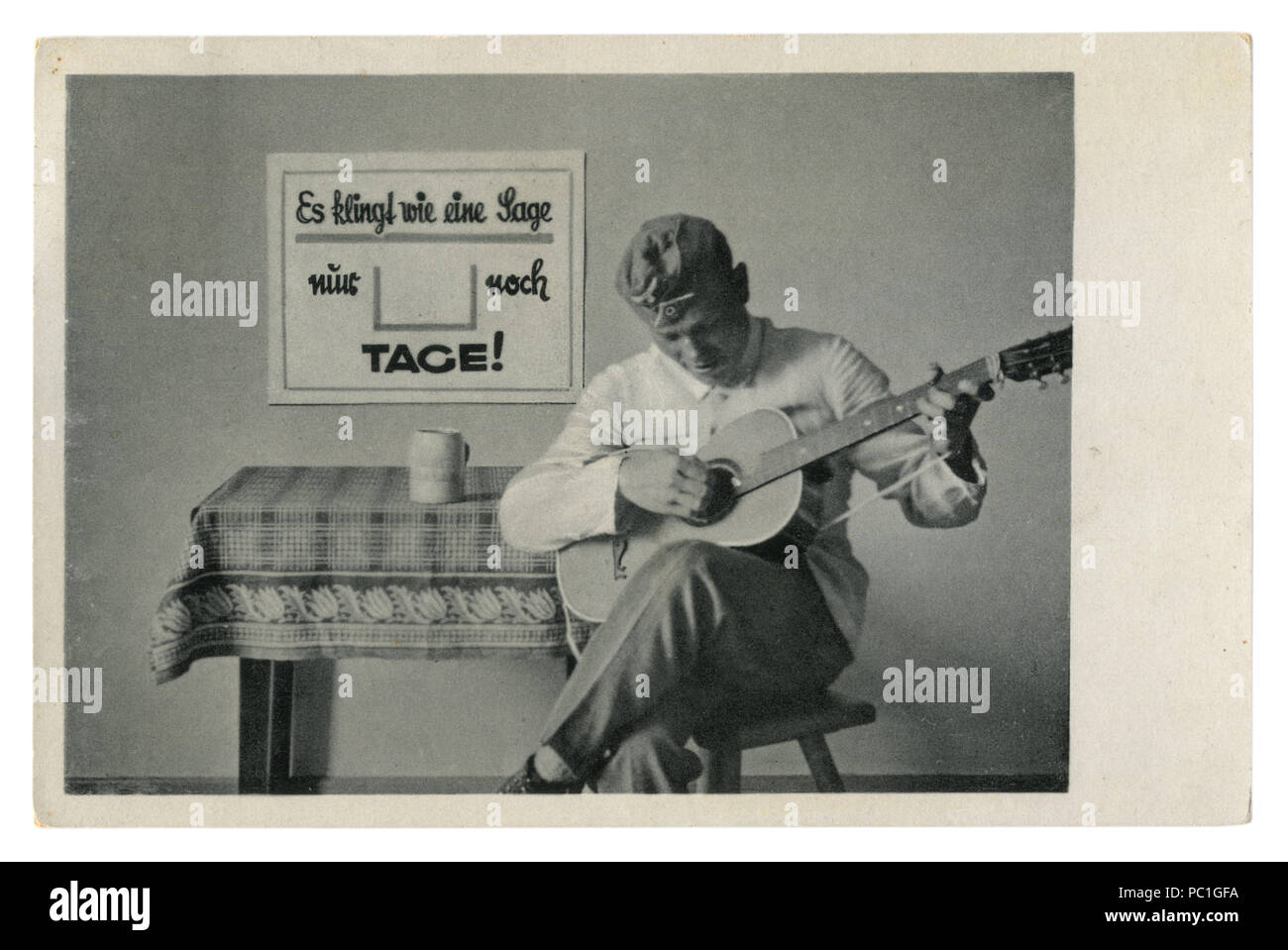German historical photo postcard: soldier in forage cap plays guitar sitting on a chair. The army's leisure, world war two, Germany, Third Reich Stock Photo