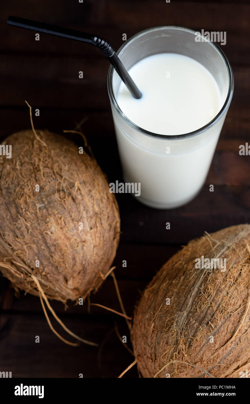 A glass of coconut milk with black straw and two whole coconuts on wooden background. Stock Photo