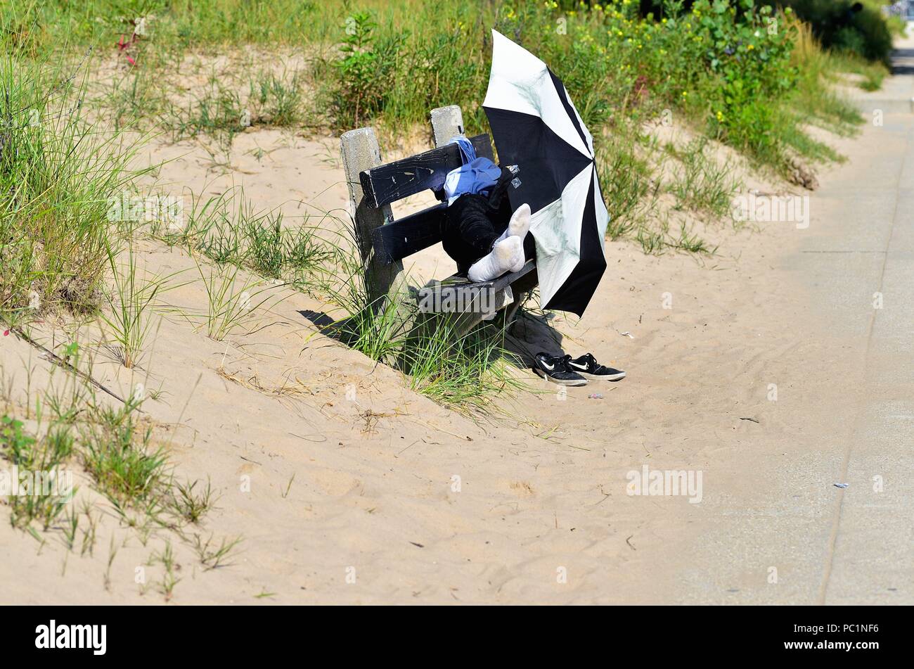 Chicago, Illinois, USA. A homeless man sleeps on a bench at Kathy Osterman Beach (also known as Hollywood Beach) using a umbrella to shield his eyes. Stock Photo