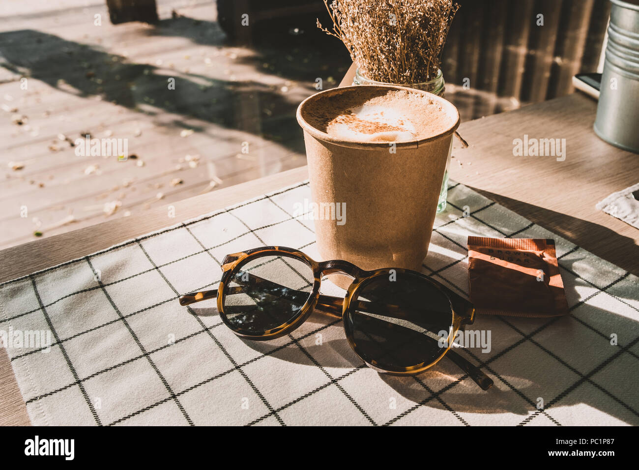 cappuccino in take away coffee cup and sunglasses on table cloth with dry flower on wood table with sunlight hard shadow at window in evening.food and Stock Photo
