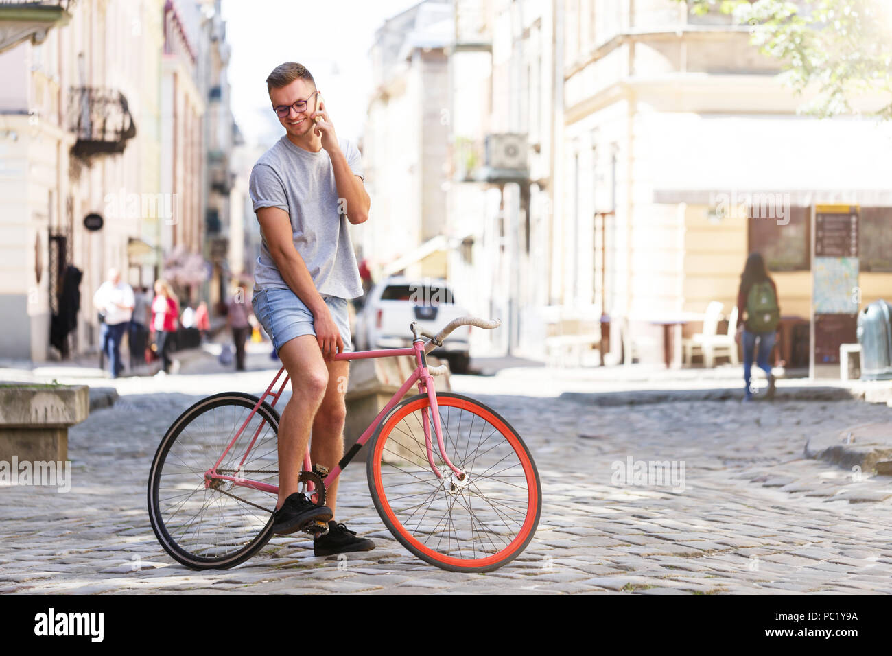 Full length portrait of handsome young caucasian man in casual clothes using phone, looking away and smiling while leaning on his bike, standing outdoors Stock Photo