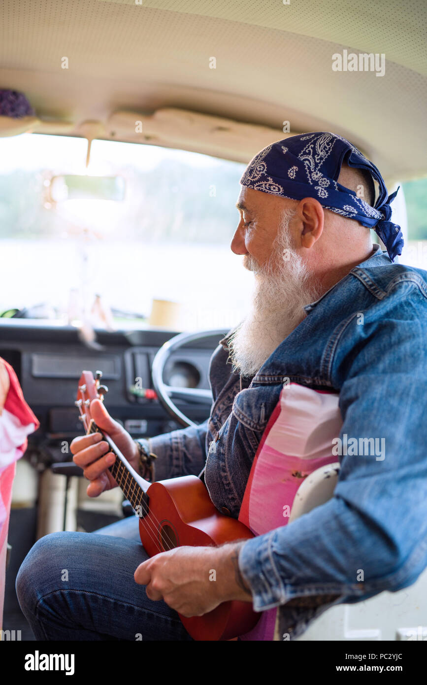 An old hipster sitting in a van playing ukulele.  Stock Photo