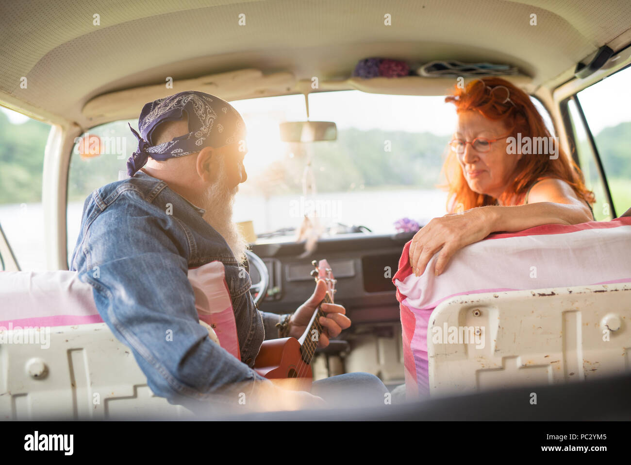 An old hipster couple sitting in a van, the man playing ukulele  Stock Photo