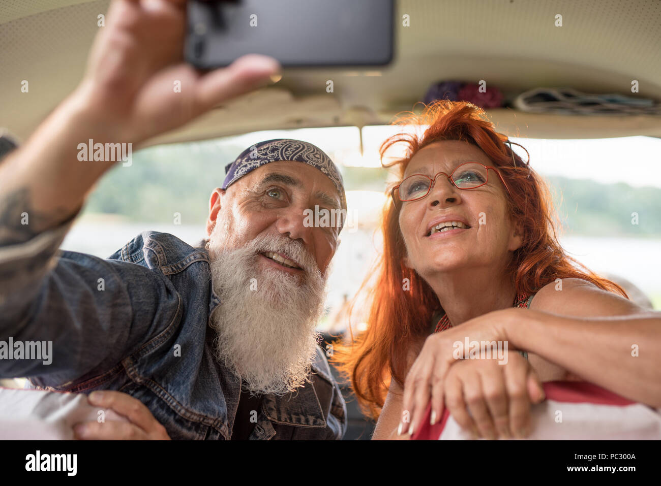 An old hipster couple sitting in a van doing a selfie with a phone. he is tattooed and he wears a white beard, she has red hair Stock Photo