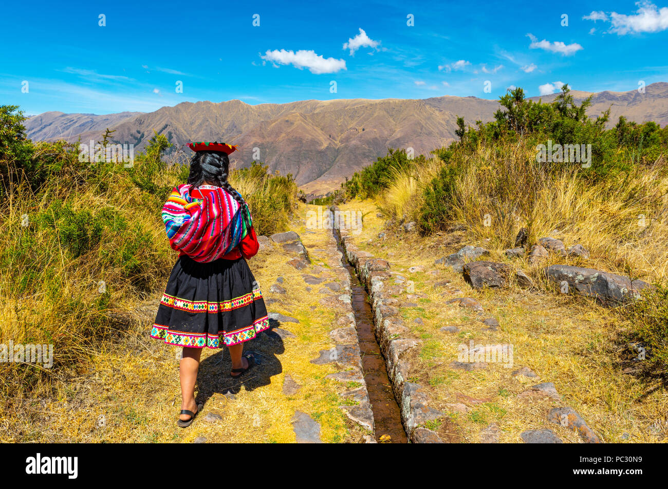 Young indigenous Quechua girl in traditional clothing walking along a aqueduct inside the Inca ruin of Tipon near the city of Cusco, Peru. Stock Photo