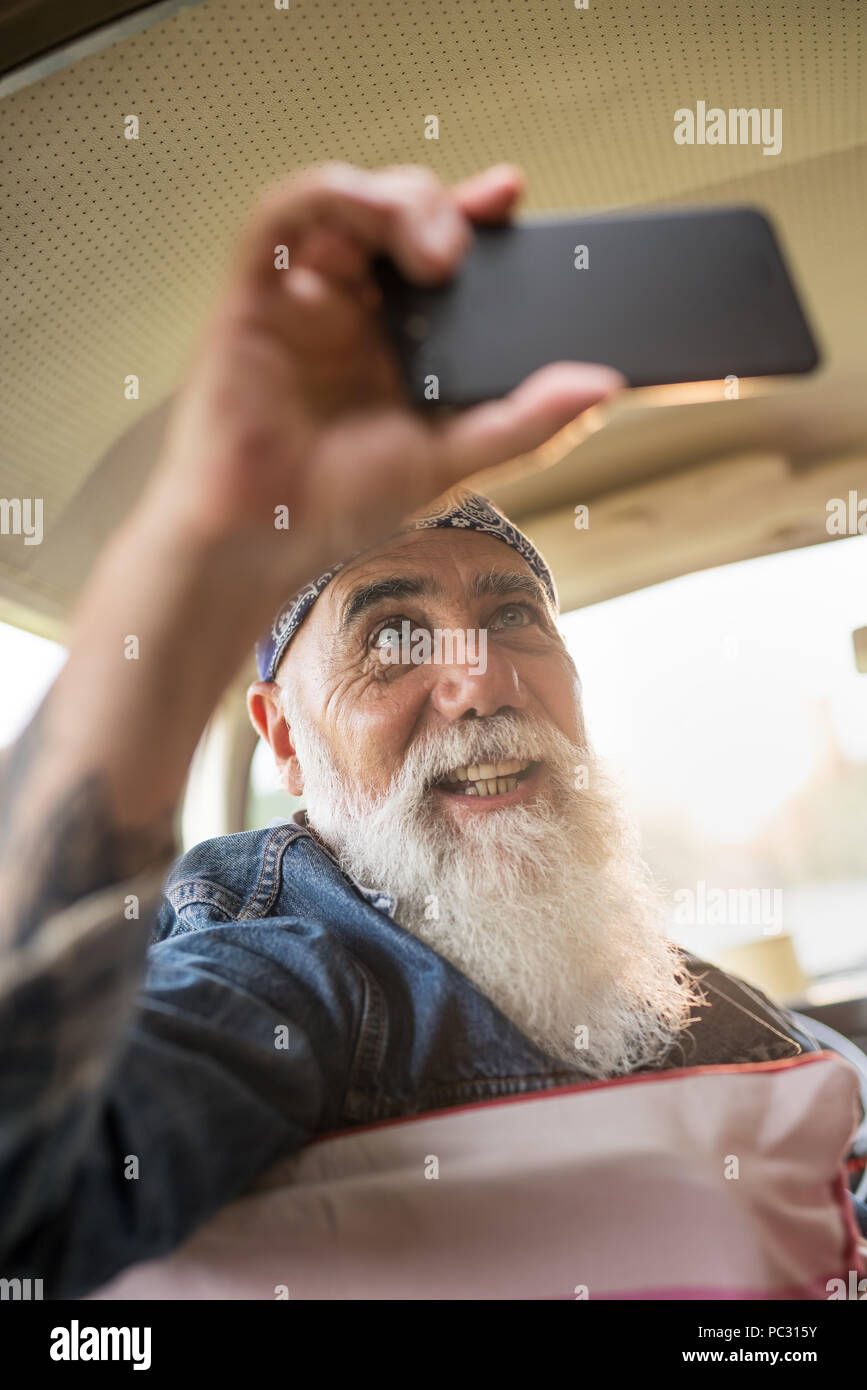 An old  hipster sitting in a car doing a selfie Stock Photo