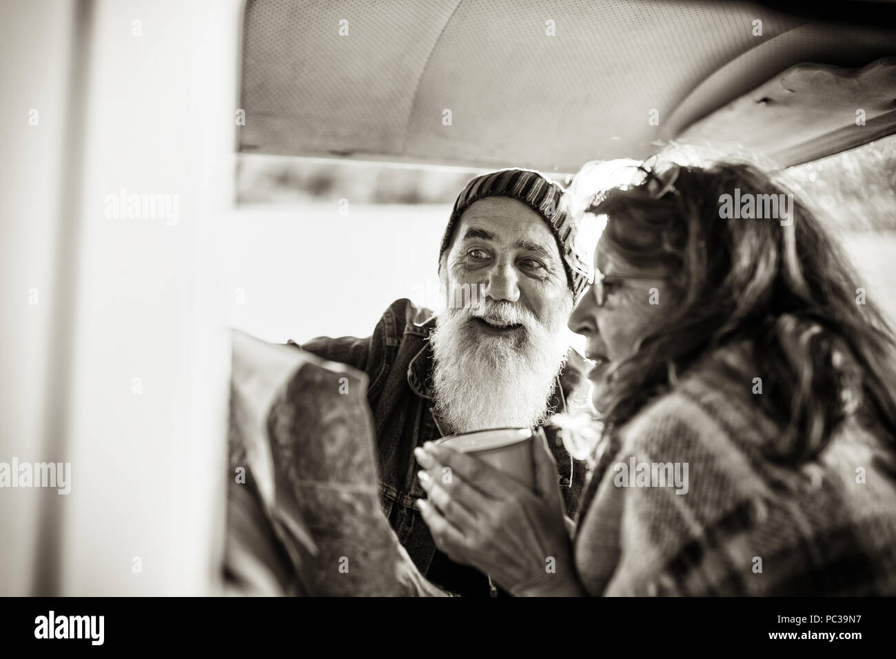 A senior hipster couple sitting in a car, drinking coffee and looking at a road map. he is tattooed and he wears a white beard. black and white Stock Photo
