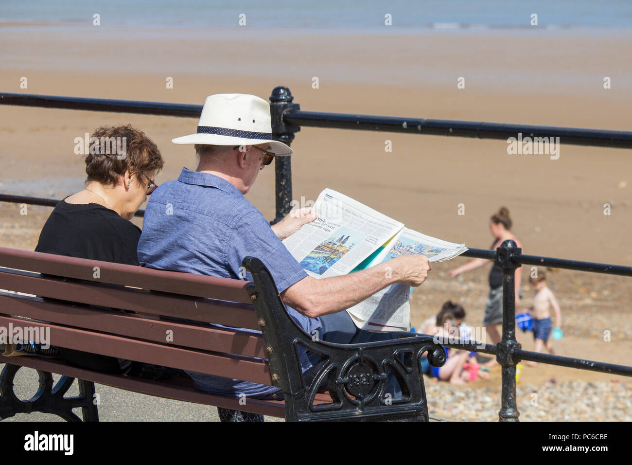 Elderly couple sitting on seat overlooking the beach. Man reading newspaper. Summer 2018. UK Stock Photo