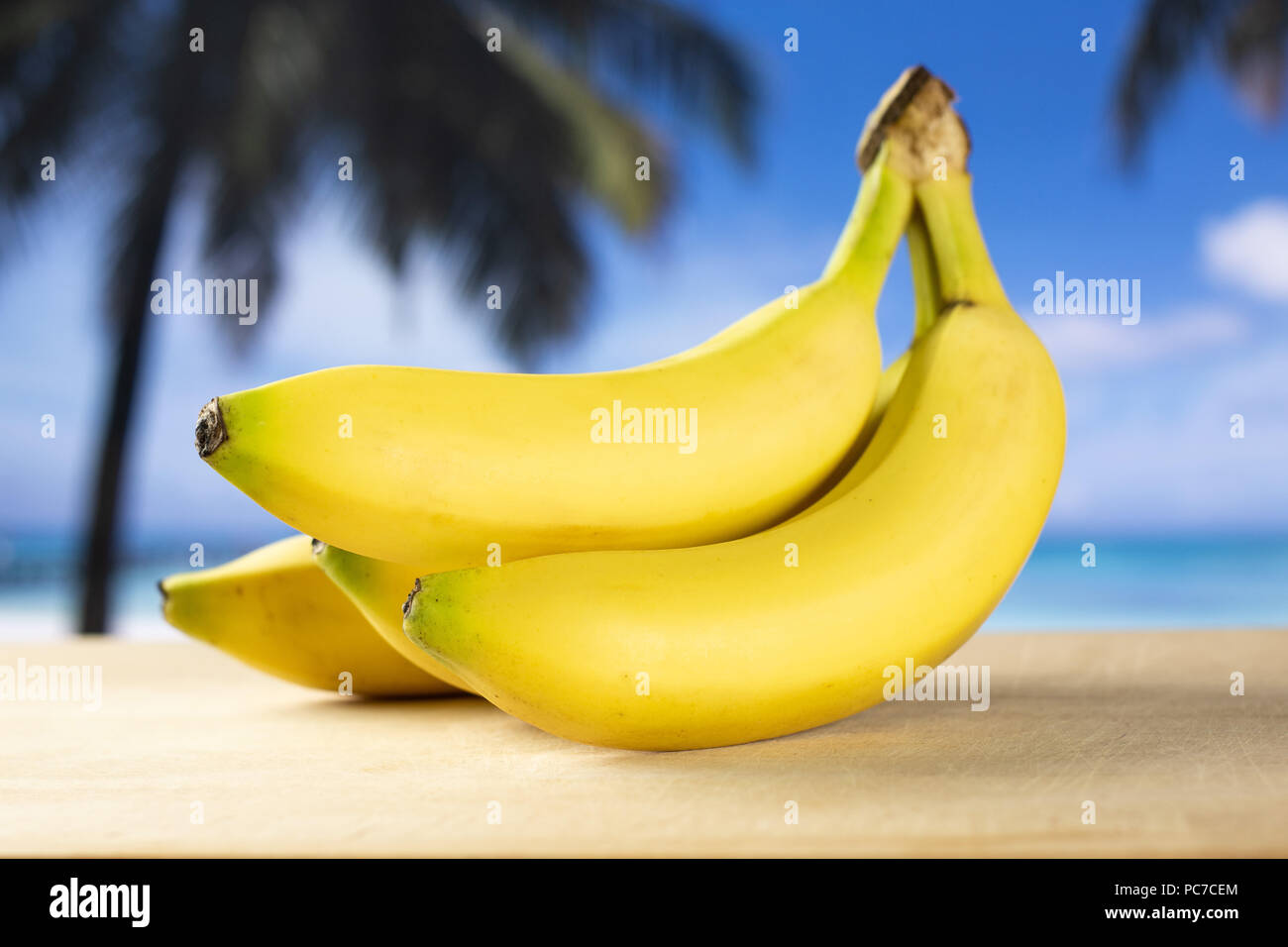 Group of three whole fresh yellow banana one cluster with palm trees on the beach in background Stock Photo