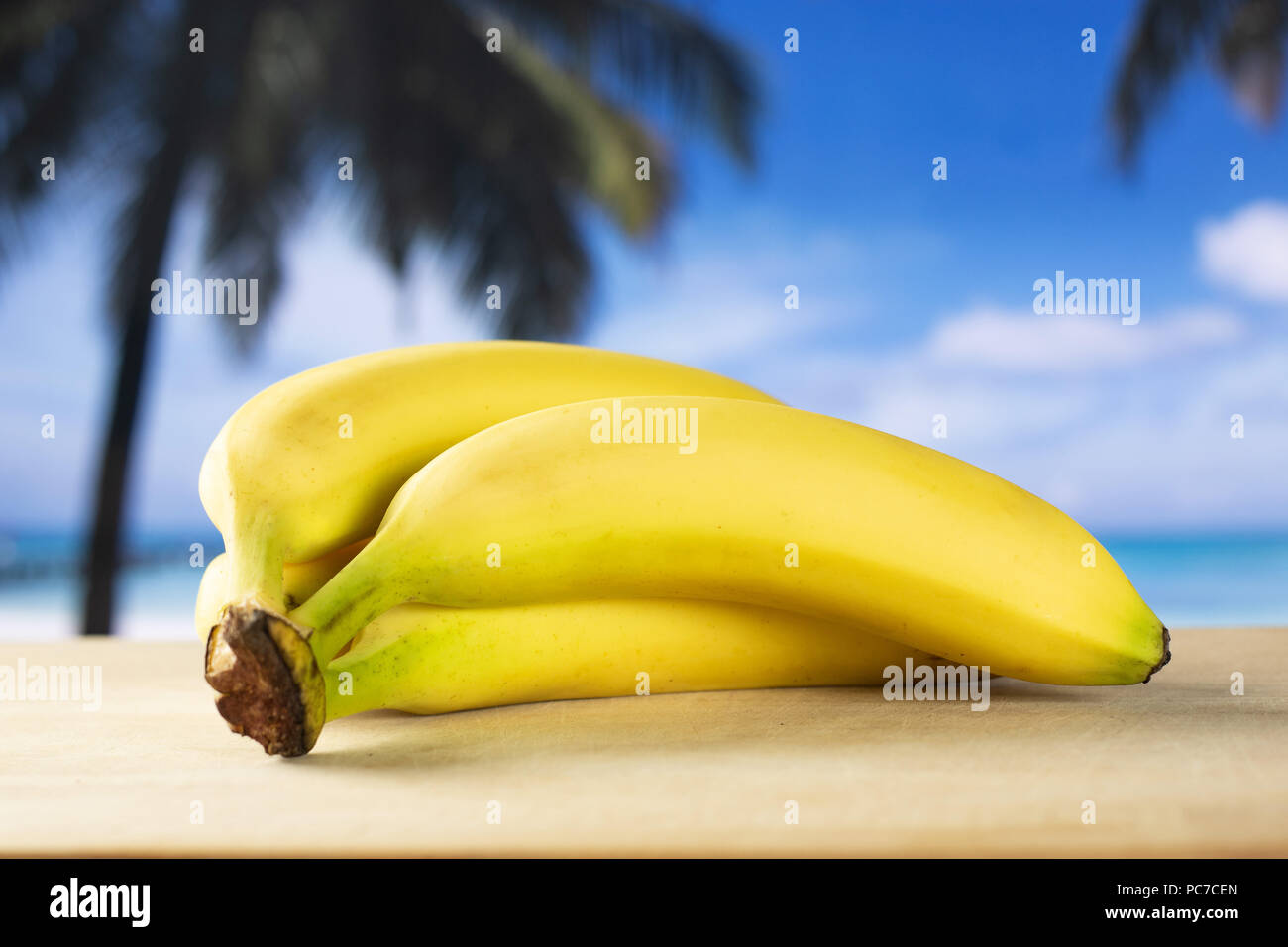 Group of four whole fresh yellow banana one ripe cluster with palm trees on the beach in background Stock Photo