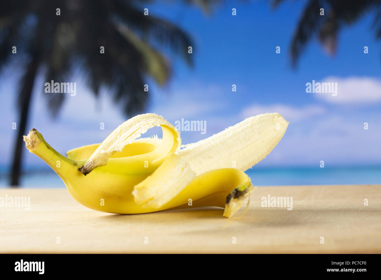 One whole open fresh yellow banana with palm trees on the beach in background Stock Photo