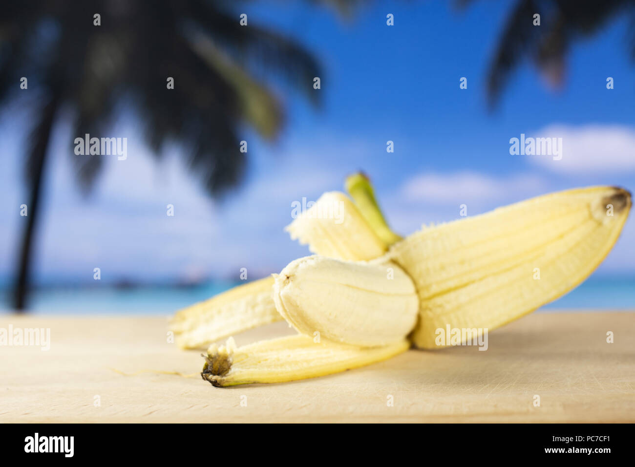 One whole fresh yellow banana opened like a plane with palm trees on the beach in background Stock Photo