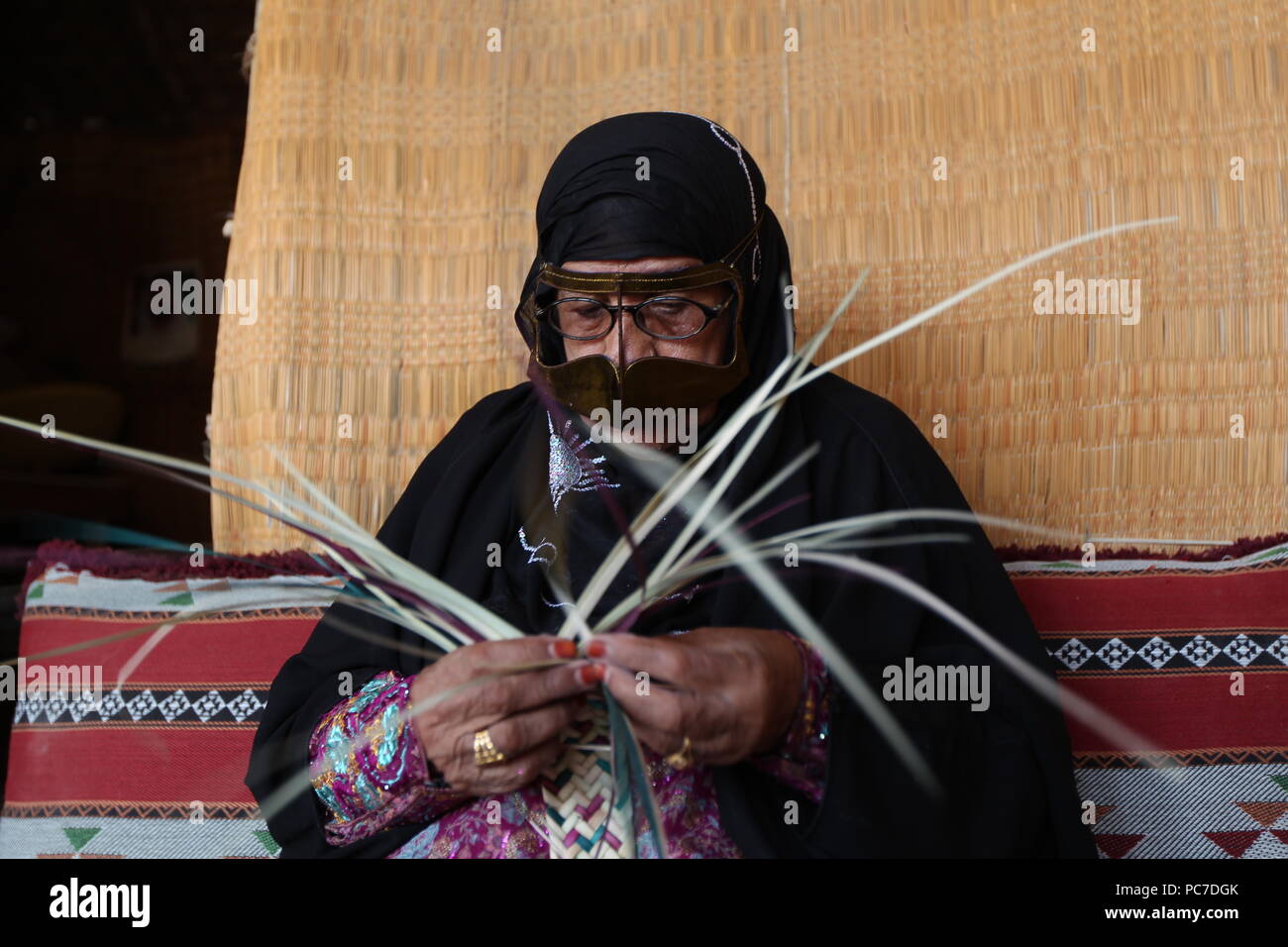 A UAE woman, sporting a metallic burqa with henna died fingers, weaves straw baskets and mats out of palm leaves at the heritage village in Fujairah. Stock Photo