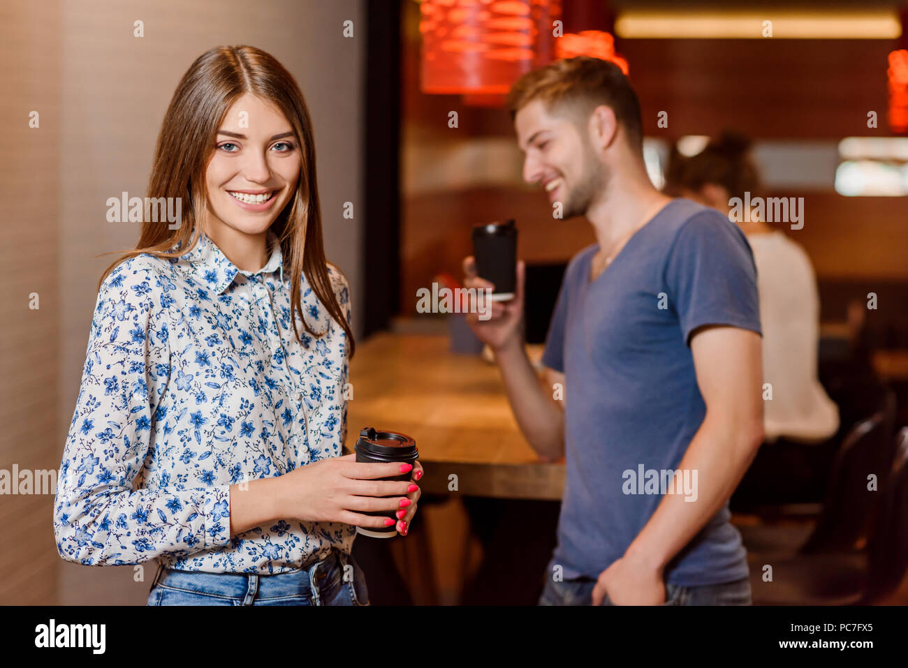 Front view of gorgeous young woman posing, looking at camera and smiling. Smiling man taking photo of his friend using phone. Positive girl holding black paper cup of coffee. Cafe interior. Stock Photo