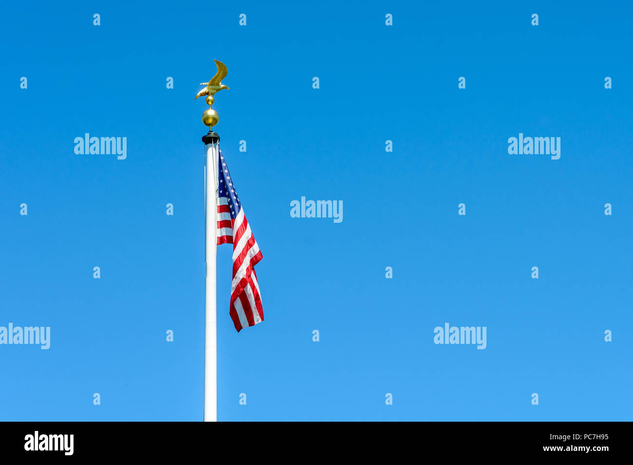 The flag of the United States of America dangling at full-mast on a white pole topped with a golden eagle on ball against blue sky. Stock Photo