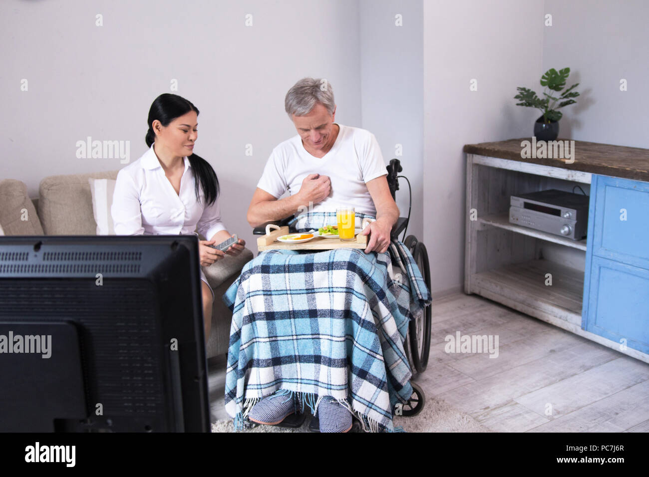 Nurse sitting next to patient with food tray. Gorgeous asian doctor sitting on couch with tv remote in her hands next to senior man in wheelchair with Stock Photo