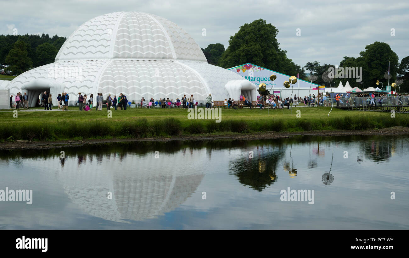 Busy showground at RHS Chatsworth Flower Show (people visiting, marquee, tents & Great Conservatory dome reflected in river) Derbyshire, England, UK. Stock Photo