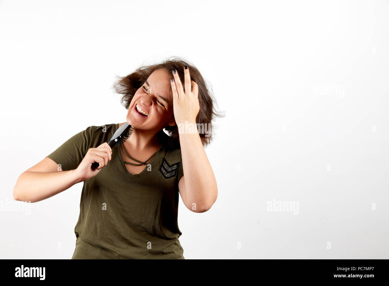 Young brunette woman with tangled unruly hair in casual khaki shirt holding comb and pressing palm to head in pain with closed eyes and open mouth sta Stock Photo