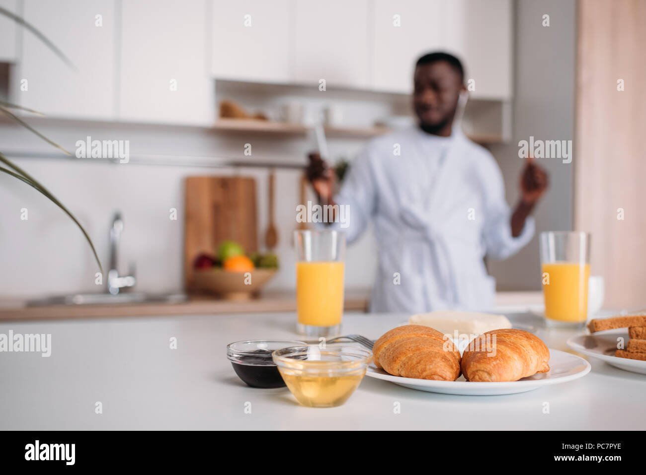 Close up view of croissants on plate, orange juice, jams and butter with young man in earphones on blurred background Stock Photo