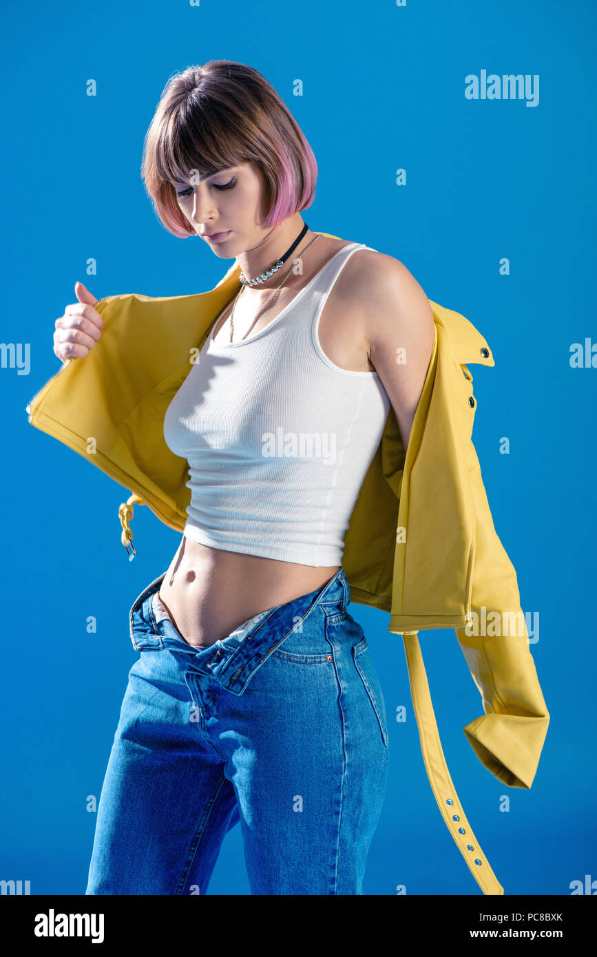 fashionable girl wearing leather jacket and standing with unbuttoned jeans isolated on blue Stock Photo