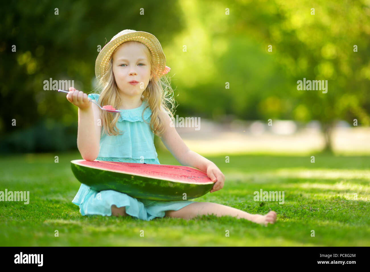 Funny little girl eating watermelon with the spoon outdoors on warm and sunny summer day. Healthy organic food for small kids. Stock Photo