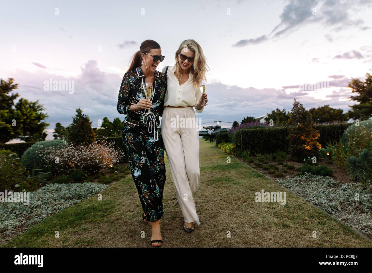 Full length of fashionable women walking together and laughing outdoors. Females friends with a glass of wine enjoying themselves outdoors. Stock Photo