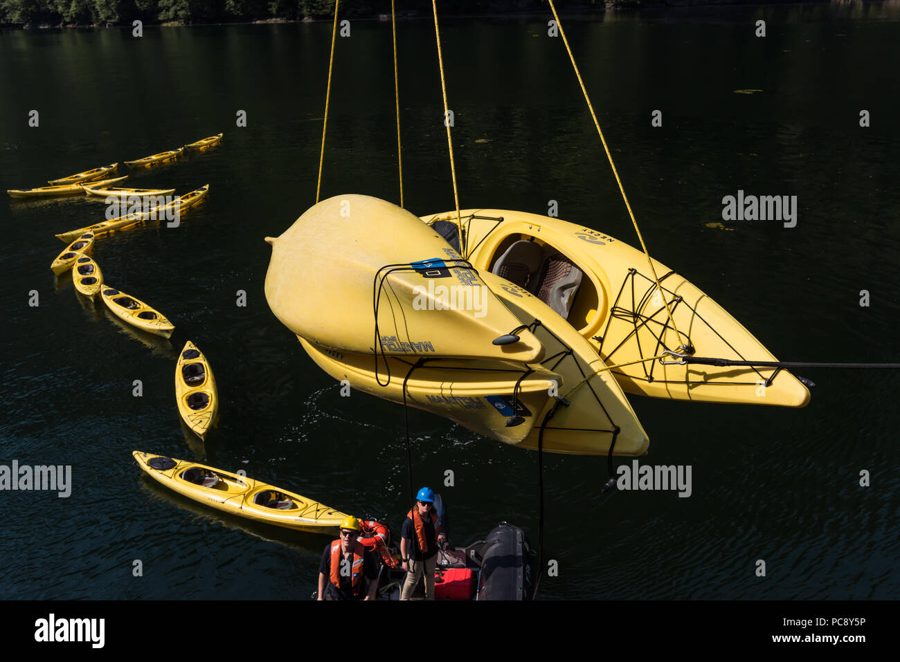 Two crew members of the National Geographic Sea Bird supervise the lowering of 4 yellow kayaks with other kayaks floating in the water behind them. Stock Photo