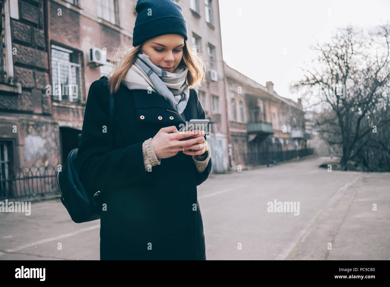 Young caucasian woman wearing wool coat, scarf and beanie using smart phone while walking on city. Female looking at mobile device on winter street. Stock Photo