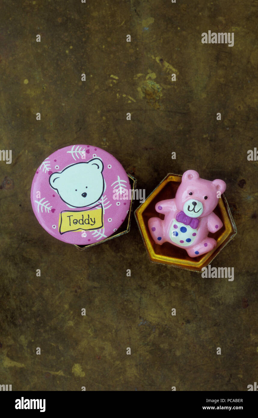 Small pink plastic teddy bear lying in hexagonal brass pill box next to pink badge with drawing of white ted labelled Teddy Stock Photo